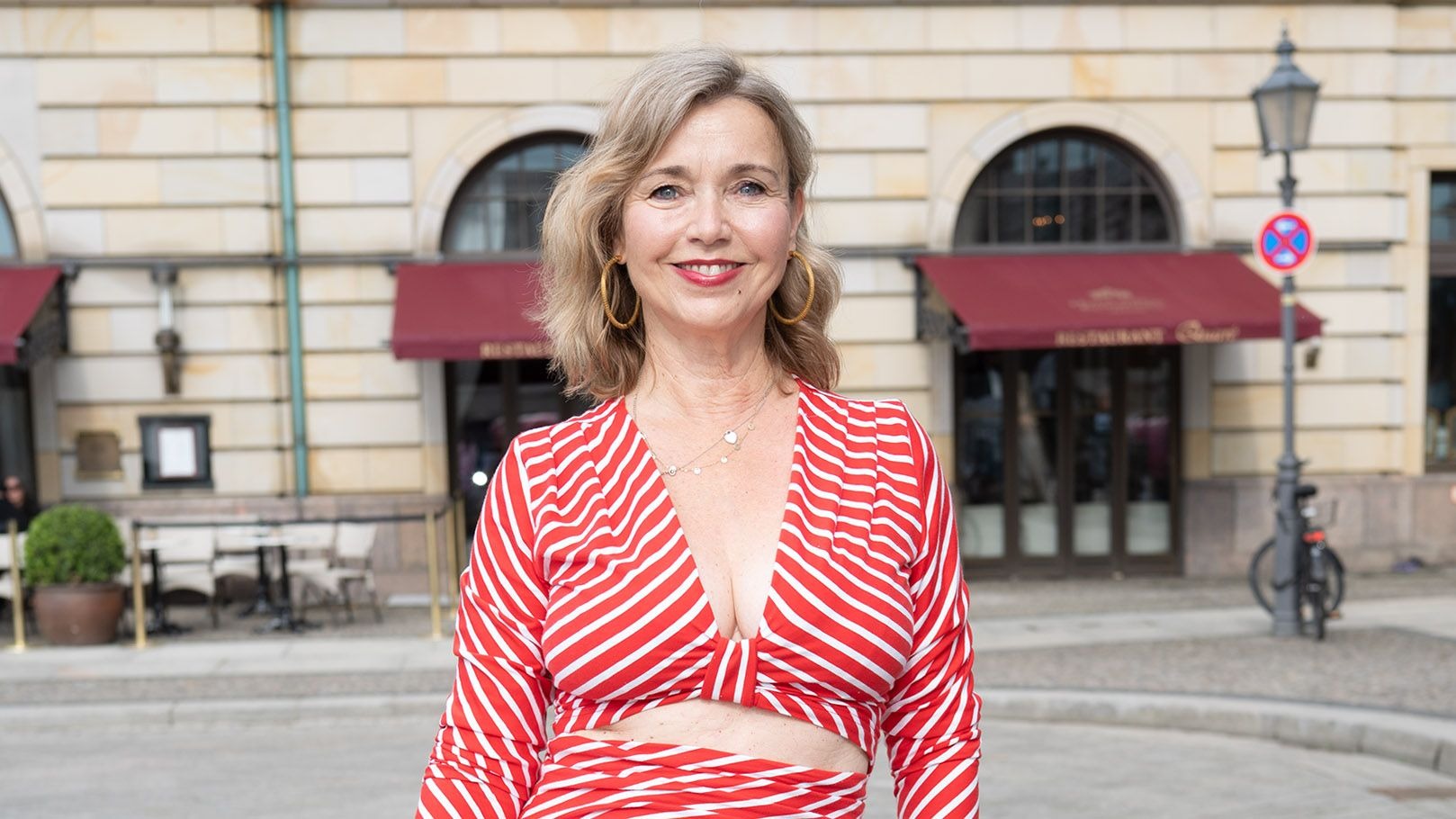 Tina Ruland lächelt in einem rot-weiß gestreiften Top, goldene Creolen tragend, vor einem Gebäude mit roten Markisen