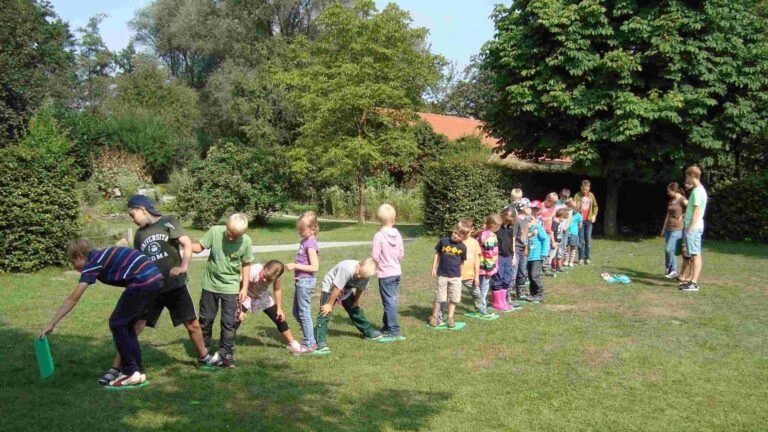 Kinder bewegen sich reihenweise auf grünen Matten über eine Wiese, umgeben von Bäumen und Sträuchern