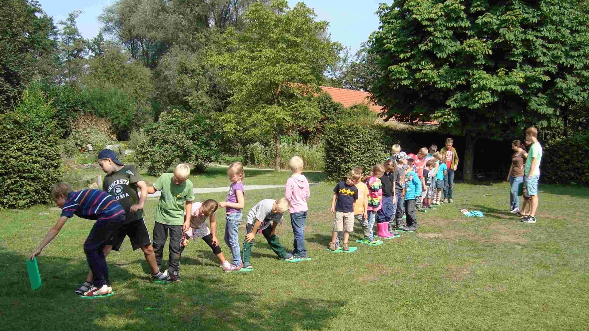 Kinder bewegen sich reihenweise auf grünen Matten über eine Wiese, umgeben von Bäumen und Sträuchern