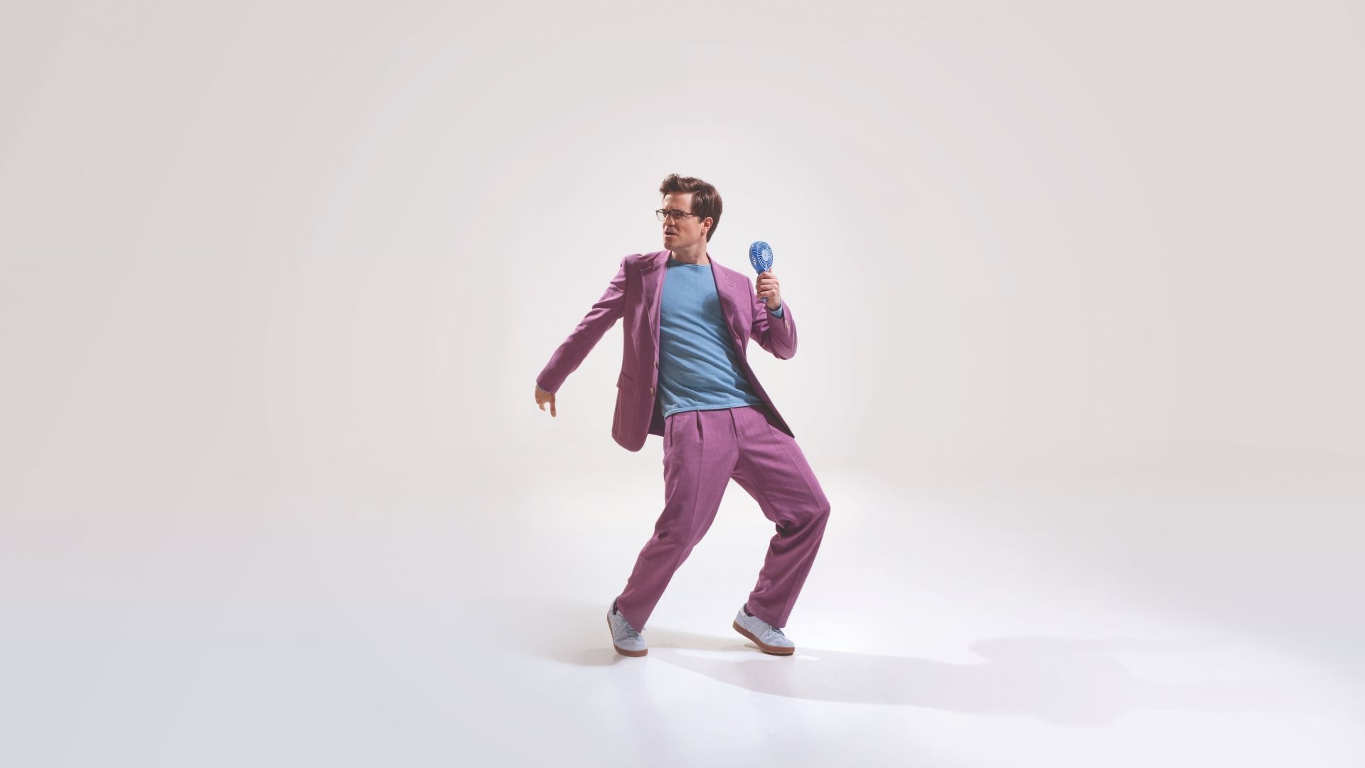 Bodo Wartke in lila Anzug und blauem Shirt hält blauen Handventilator in dynamischer Pose auf hellem Grund