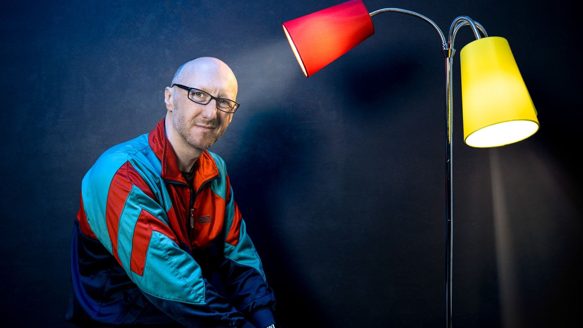 Ralf Senkel mit Glatze und Brille im bunten Trainingsanzug lächelt vor dunklem Hintergrund neben einer Stehlampe