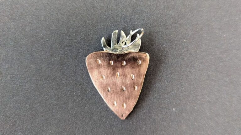 Erdbeerförmiger Metallanhänger mit kleinen Vertiefungen und silbernen Blättern auf dunklem Untergrund