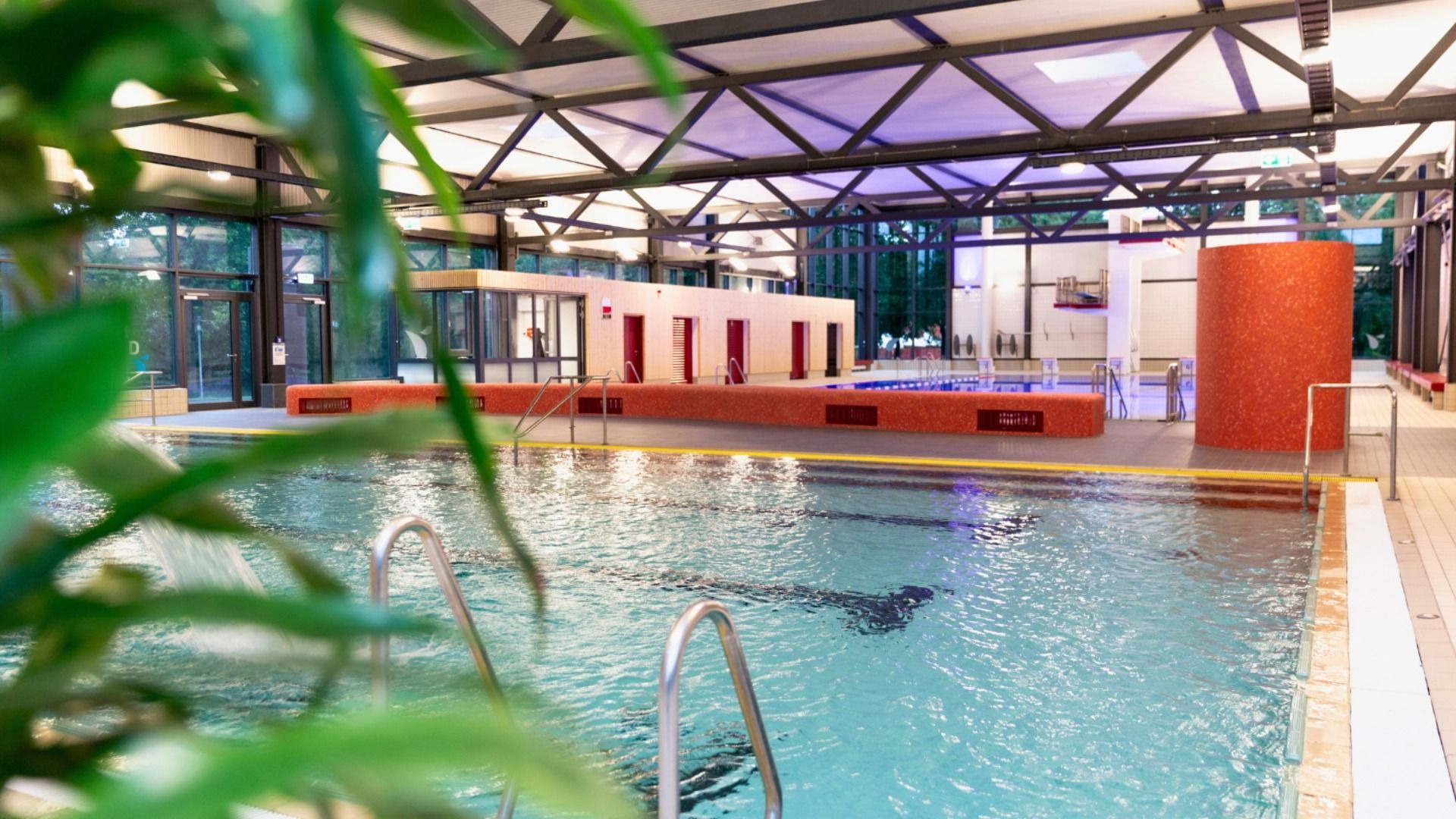 Großes, helles Hallenbad mit klarem Wasser, Edelstahlleitern, roten Kabinen und einer Sprunganlage im Hintergrund