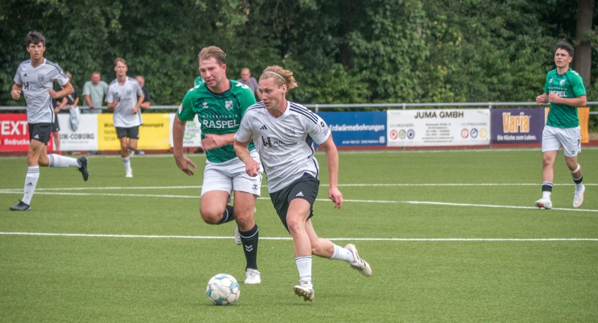 Zwei Fußballspieler in hellgrauem und grünem Trikot rennen mit Ball auf einem grünen Rasenfeld, weitere Spieler im