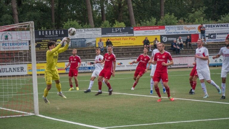 Ein Fußballtorwart in gelbem Trikot springt nach dem Ball vor dem Tor, umgeben von Spielern in roten und weißen Trikots