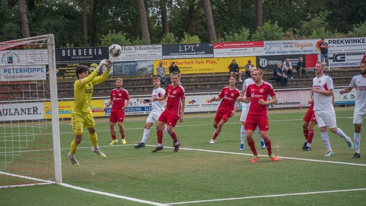 Ein Fußballtorwart in gelbem Trikot springt nach dem Ball vor dem Tor, umgeben von Spielern in roten und weißen Trikots