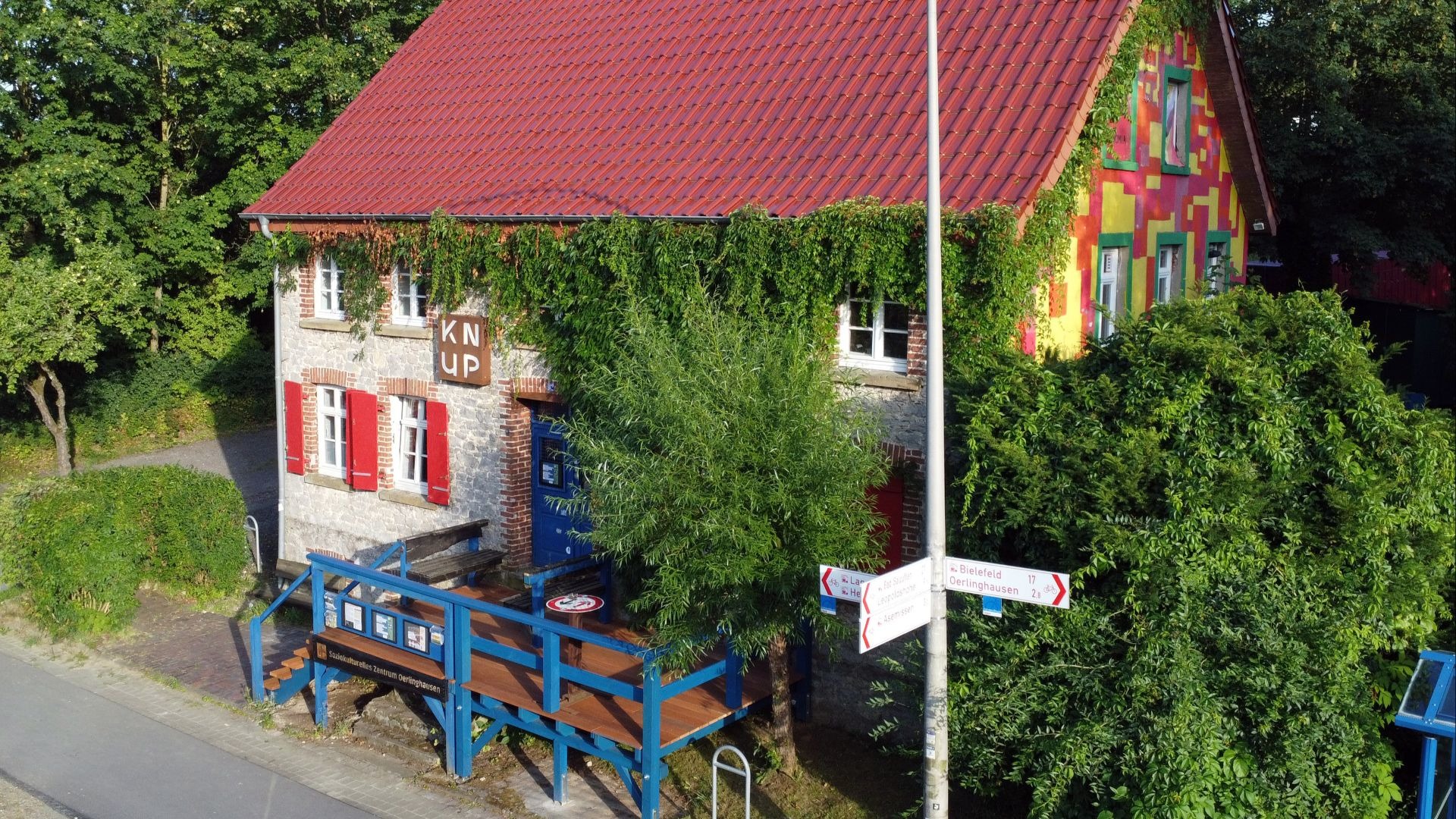 Zweigeteiltes Haus mit rotem Dach, eine Seite aus grauem Stein mit Efeu, die andere bunt bemalt, davor blaue Terrasse