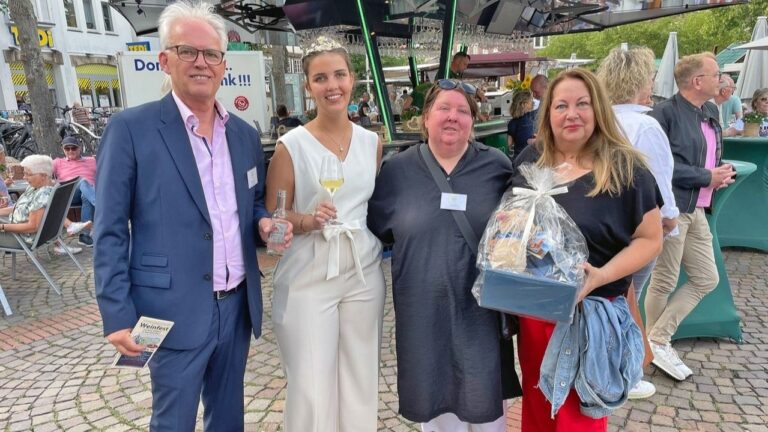 Vier Personen auf einem Weinfest: Ein Mann hält einen Flyer, eine Frau ein Weinglas, eine weitere Frau einen Präsentkorb