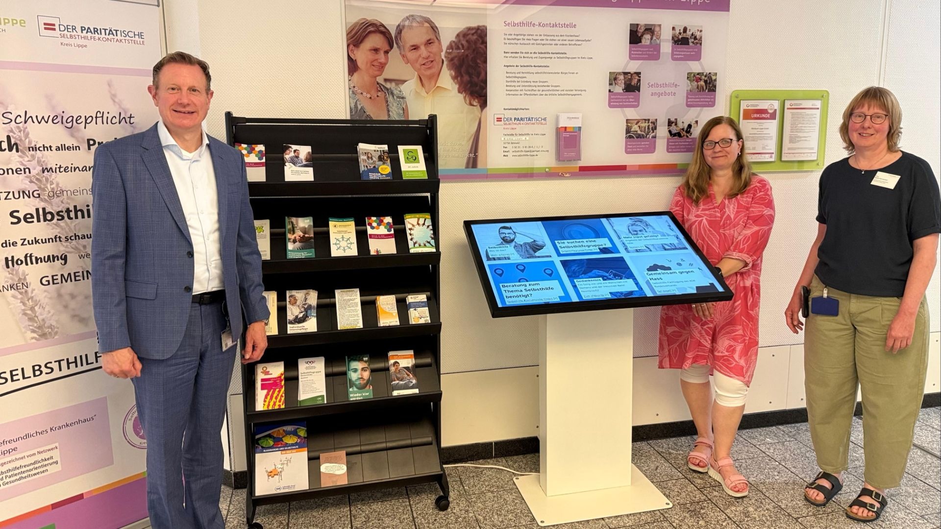 Ein Mann und zwei Frauen stehen lächelnd vor einem Infostand und einem digitalen Terminal zu Selbsthilfegruppen im