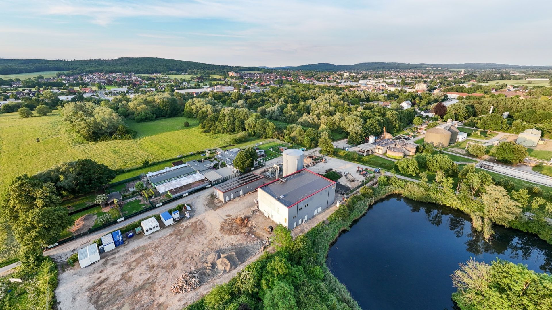 Luftaufnahme eines Industriekomplexes mit großen Gebäuden und Silos an einem dunklen Gewässer, umgeben von grüner