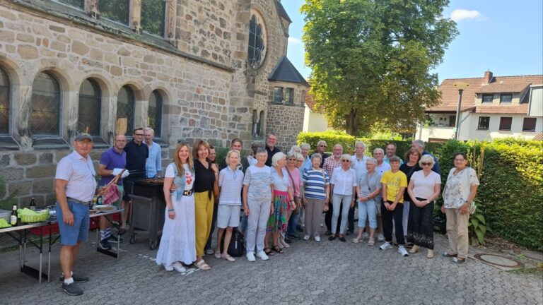 Viele Menschen stehen lächelnd vor einem Steingebäude, ein Mann grillt Würstchen an einem Grill neben Tischen mit