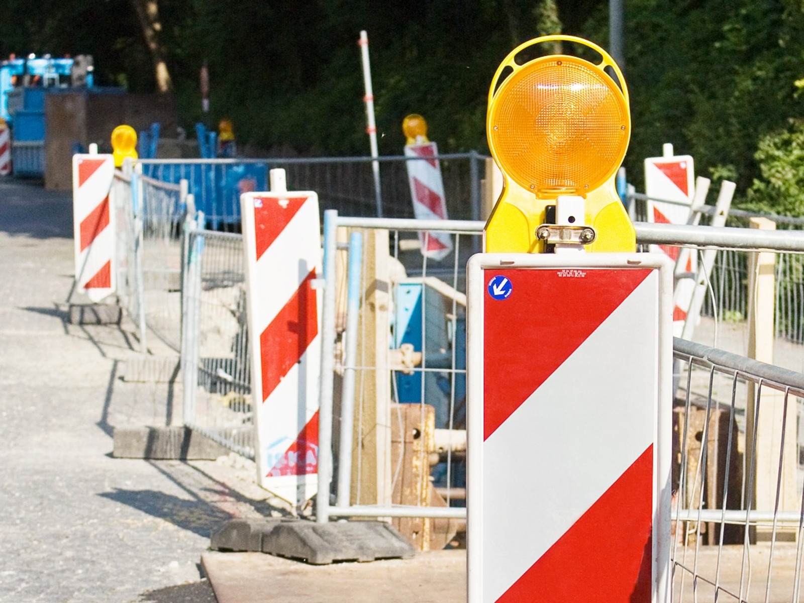 Baustelle mit rot-weißem Warnschild und gelbem Blinklicht, flankiert von Bauzäunen und weiteren Baken