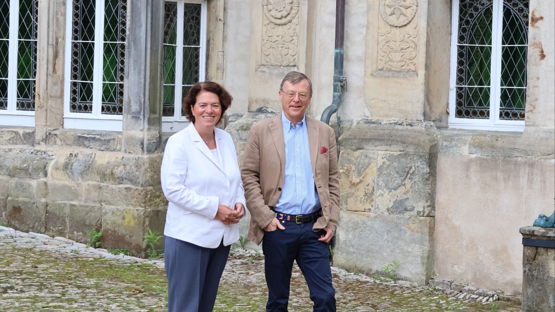 Kerstin Vieregge und ein Mann stehen lächelnd vor einem historischen Steingebäude auf gepflastertem Hof
