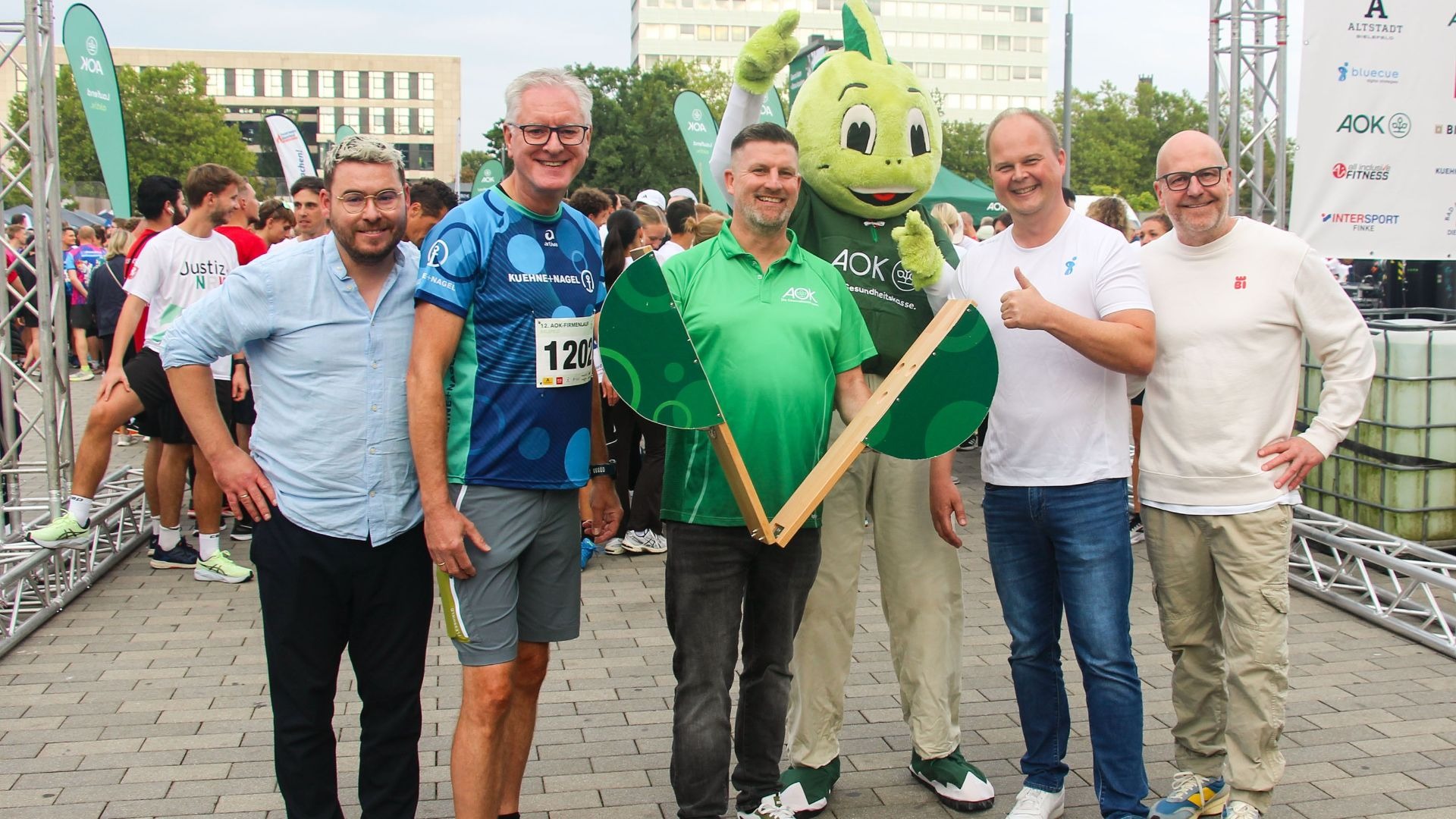 Fünf Männer und ein grünes AOK-Maskottchen lächeln beim AOK-Firmenlauf Bielefeld, einer hält ein grünes Holzelement