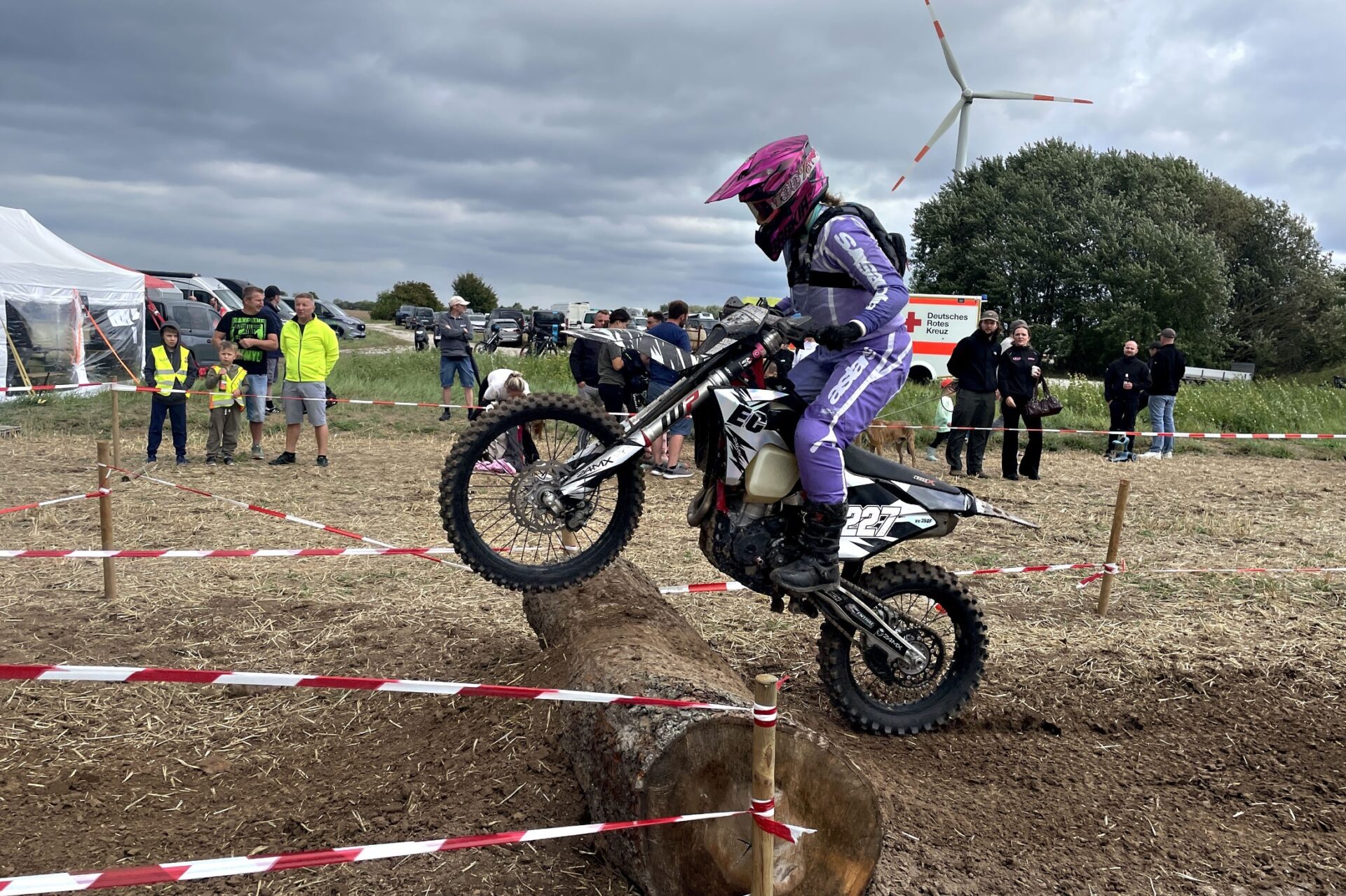 Fahrer in lila Enduro-Ausrüstung überquert auf einem Motorrad einen Baumstamm auf der Rennstrecke vor Zuschauern