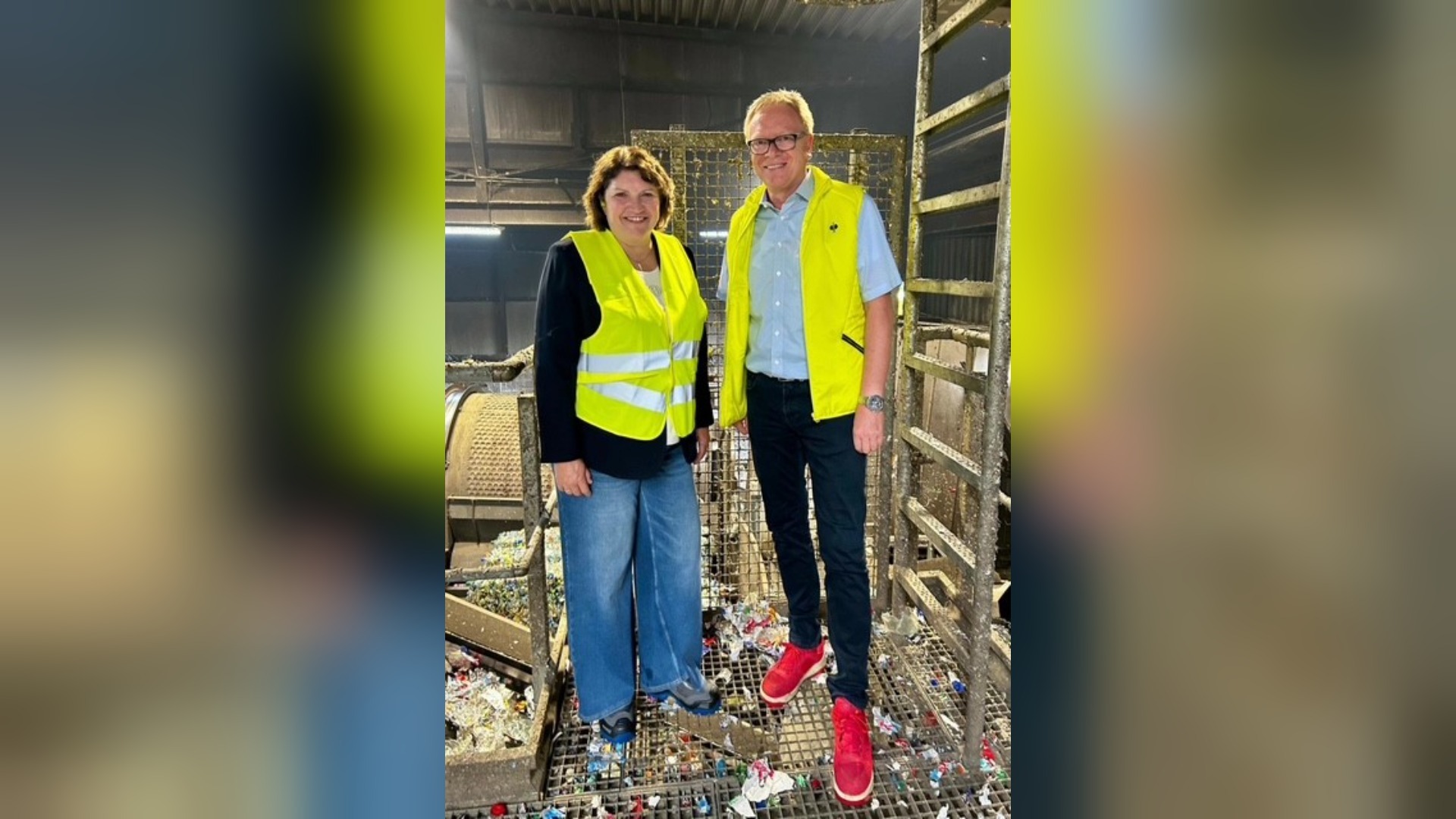 Eine Frau und ein Mann in gelben Warnwesten stehen in einer Recyclinghalle auf einem Gitterboden mit PET-Material