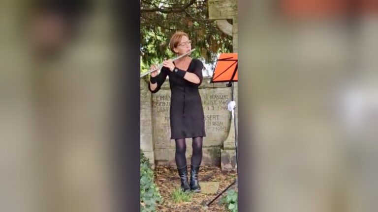 Frau in schwarzem Kleid spielt Querflöte vor einer beschrifteten Steinwand mit Notenständer