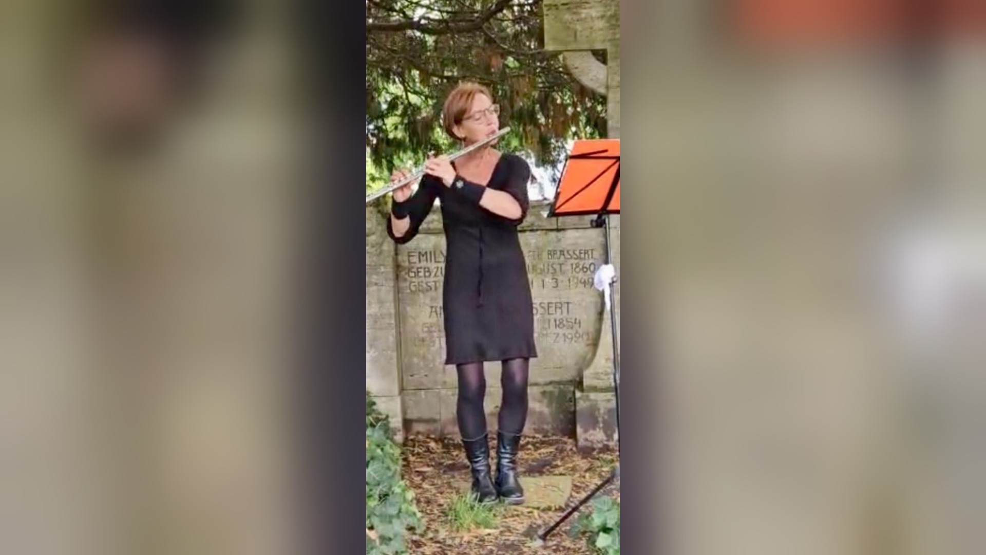 Frau in schwarzem Kleid spielt Querflöte vor einer beschrifteten Steinwand mit Notenständer