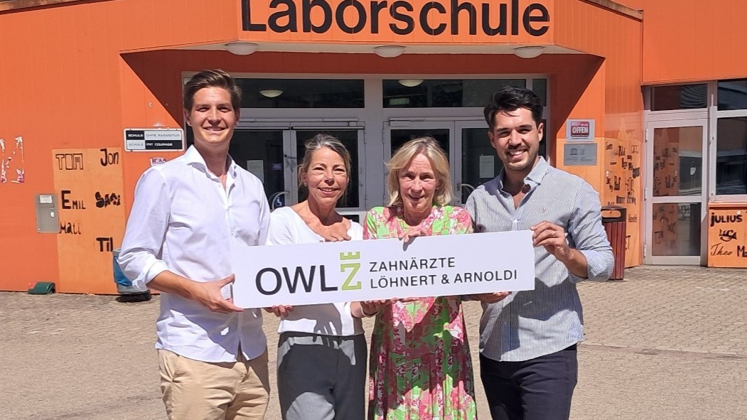 Vier lächelnde Personen halten ein Schild mit "OWL Z Zahnärzte Löhnert & Arnoldi" vor einem orangefarbenen Gebäude