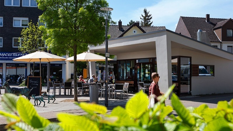 Modernes Café mit Gästen unter Sonnenschirmen auf einem Platz, davor eine Ziegenskulptur und eine Person geht vorbei