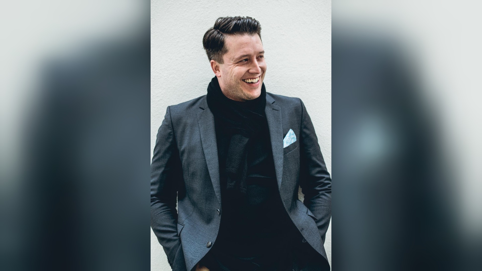 Junger lächelnder Mann im dunklen Anzug mit schwarzem Schal und blauem Einstecktuch lehnt an einer hellen Wand