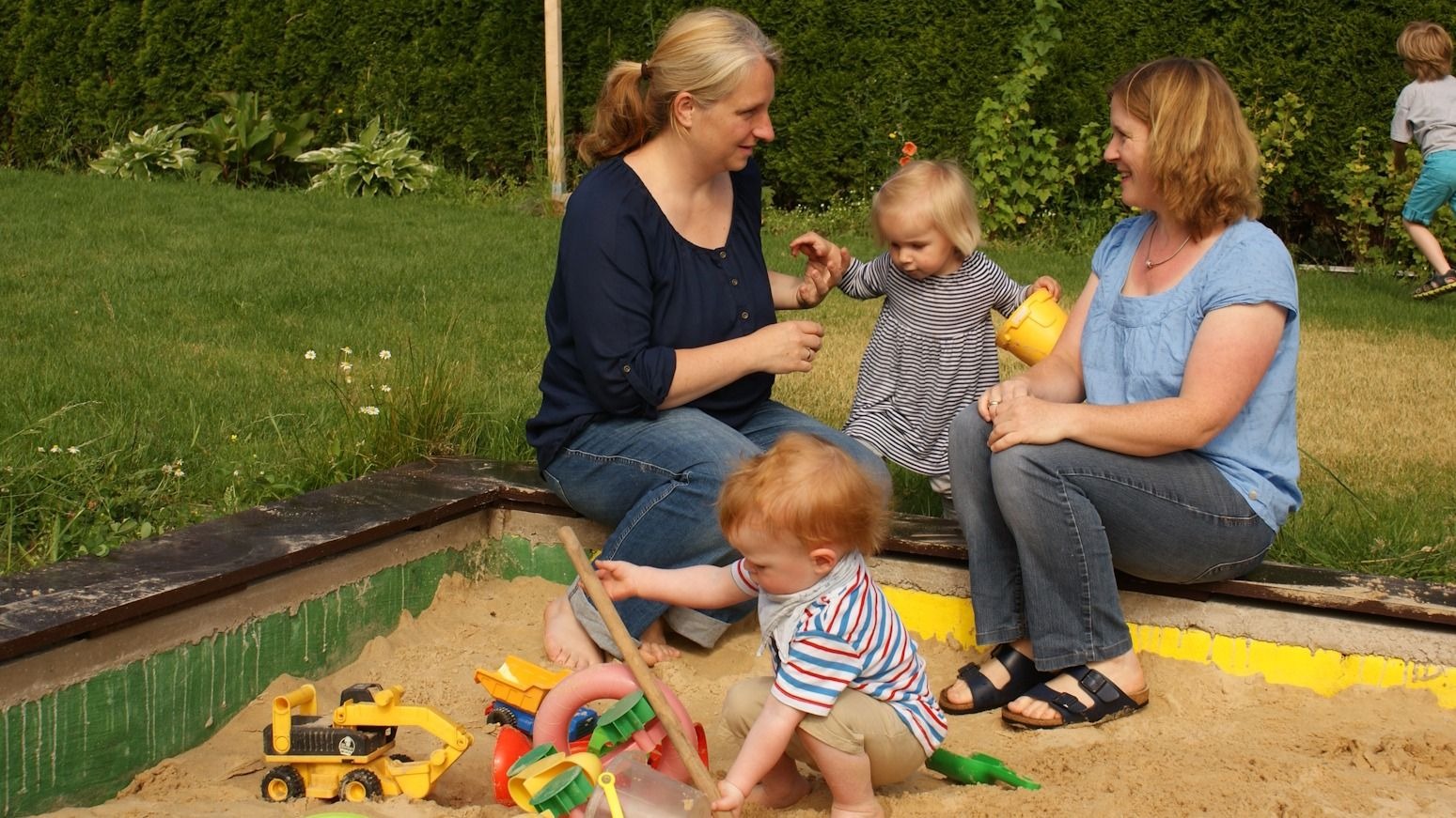 Zwei Frauen sitzen am Sandkasten und beobachten zwei Kleinkinder beim Spielen, ein weiteres Kind im Hintergrund