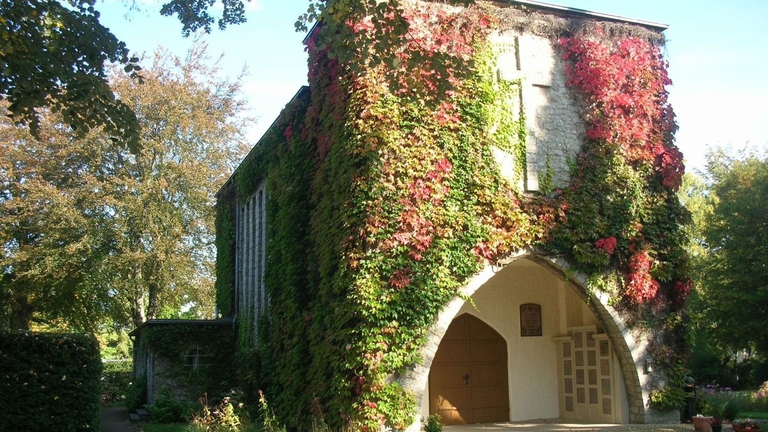 Friedhofskapelle Sieker mit Bogenportal und Holztür, stark mit grünem und rotem Efeu bewachsen, umgeben von Bäumen