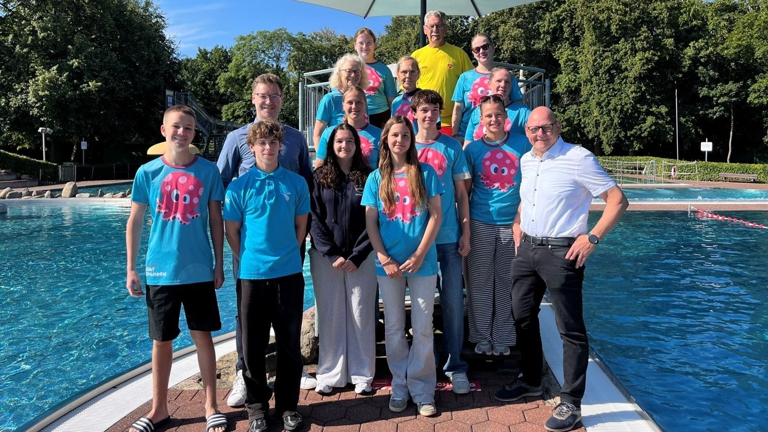 Gruppe von 16 Personen, viele in blauen T-Shirts mit Oktopus-Logo, stehen lächelnd am Schwimmbecken