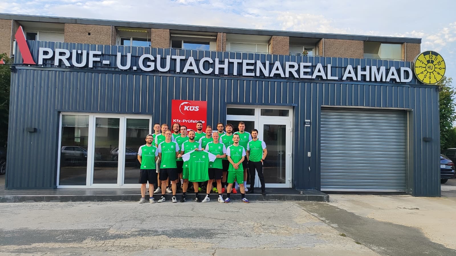 Gruppe Männer in grünen Trikots hält ein Trikot vor dem Gebäude „PRÜF- U. GUTACHTENAREAL AHMAD“