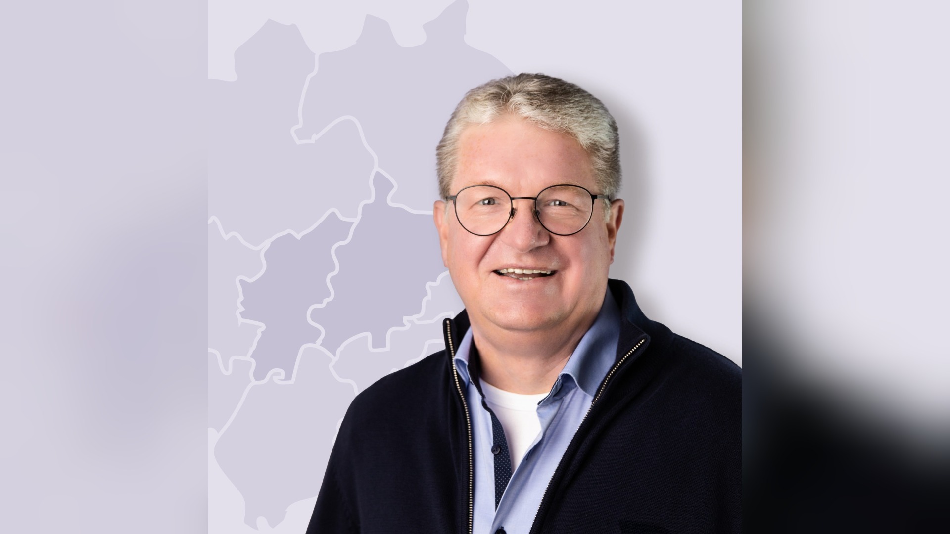Frank Sommerfeld, ein lächelnder Mann mit grauen Haaren und Brille, in blauem Hemd und Pullover vor einer Karte