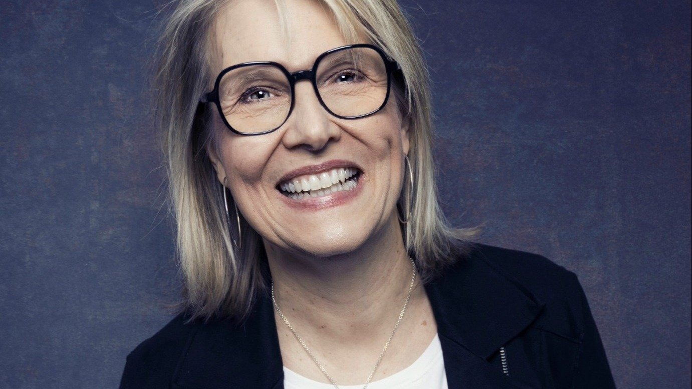 Blonde Frau Sturm mit Brille und Ohrringen lacht breit und zeigt ihre Zähne vor dunklem Hintergrund