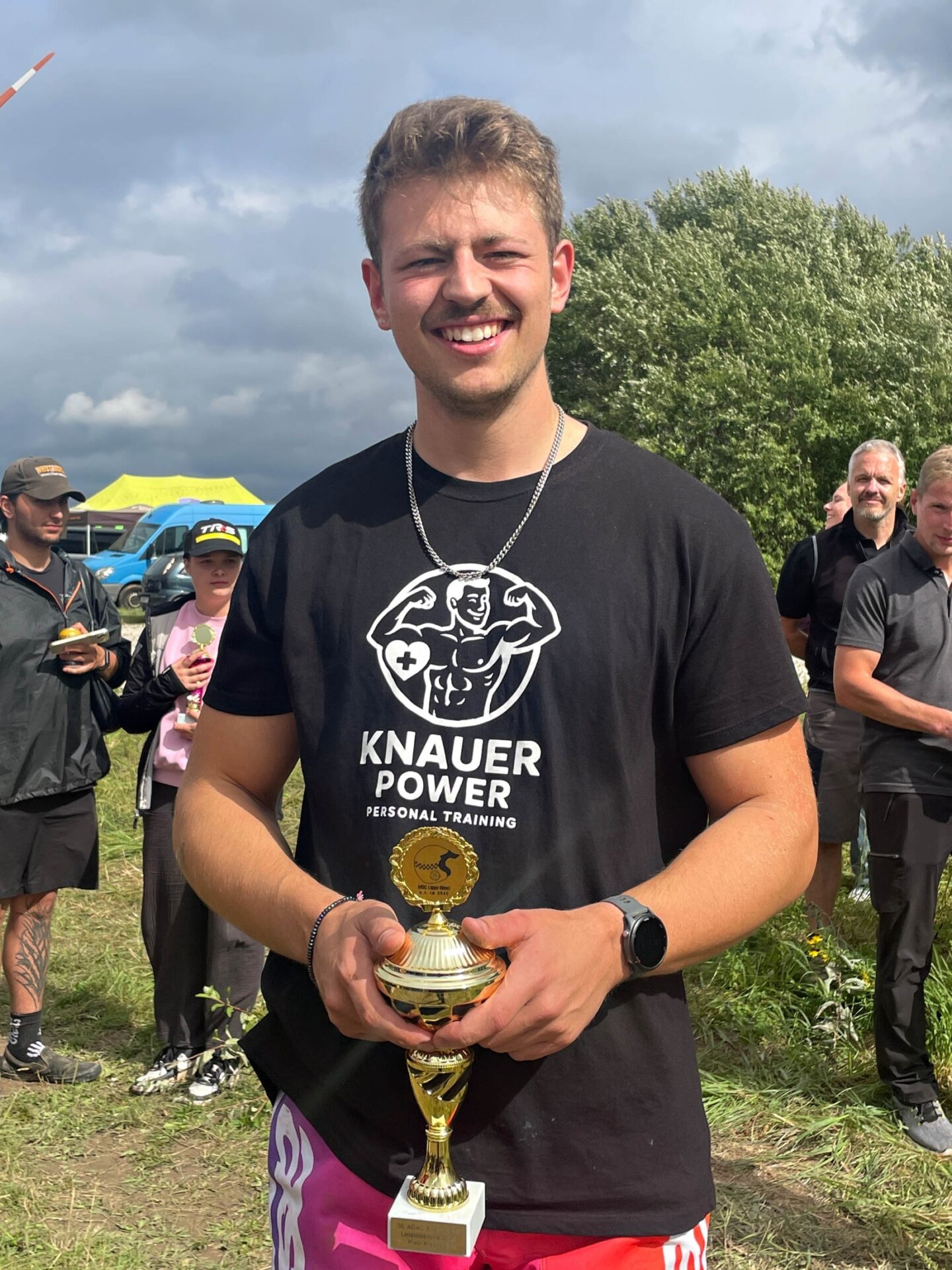 Niels Knauer lächelt, hält einen goldenen Pokal und steht im Freien vor anderen Personen