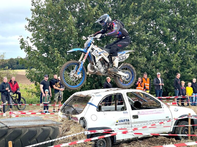 Motorradfahrer springt mit blauem Dirtbike über ein weißes, kaputtes Auto vor Zuschauern und Bäumen