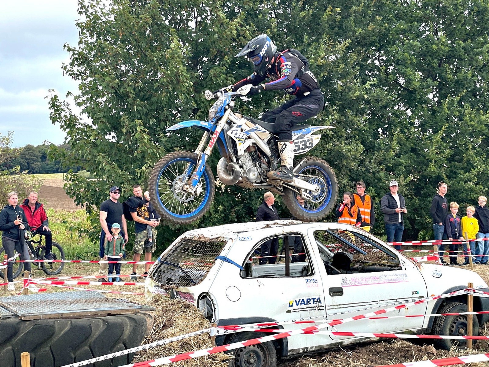 Motorradfahrer springt mit blauem Dirtbike über ein weißes, kaputtes Auto vor Zuschauern und Bäumen
