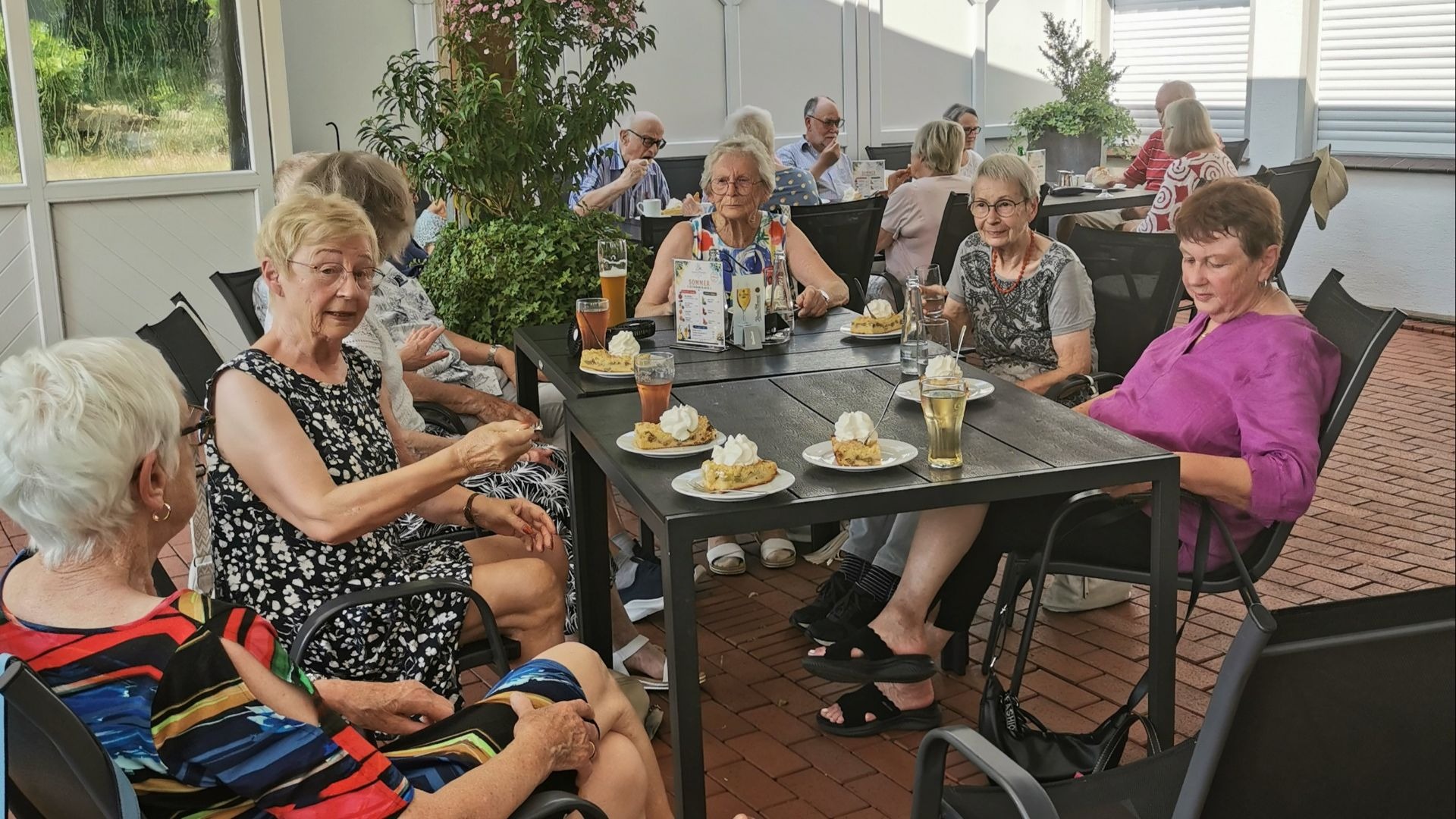 Eine Gruppe Senioren sitzt auf einer überdachten Terrasse an Tischen mit Kuchen und Getränken