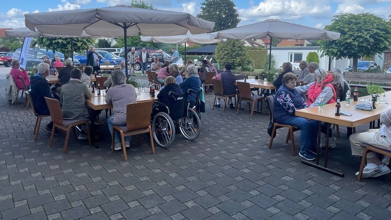 VdK-Mitglieder, teils im Rollstuhl, versammeln sich unter Sonnenschirmen an Holztischen für ein Grillfest im Freien