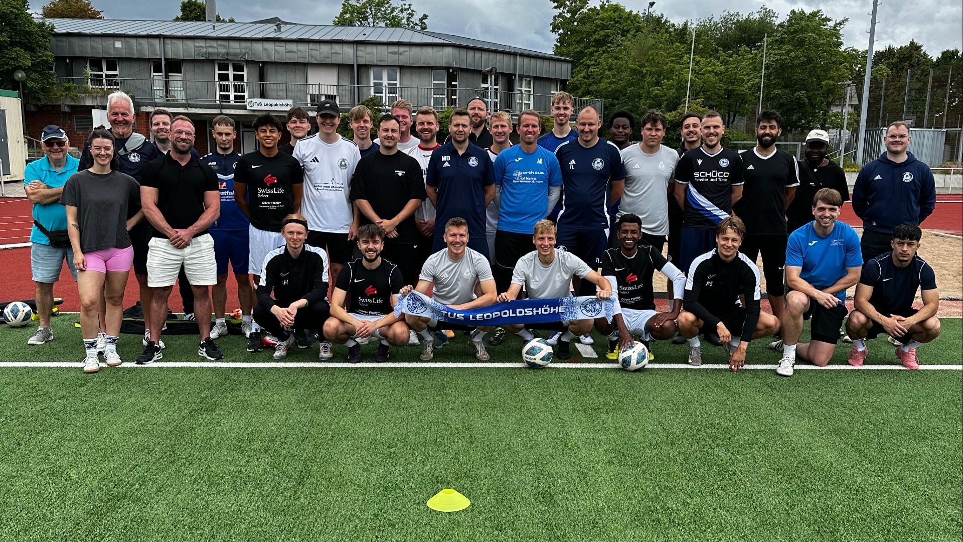 Große Gruppe von Personen posiert auf einem Kunstrasen-Sportplatz, einige halten ein blaues Banner "FÜR LEOPOLDSHÖHE