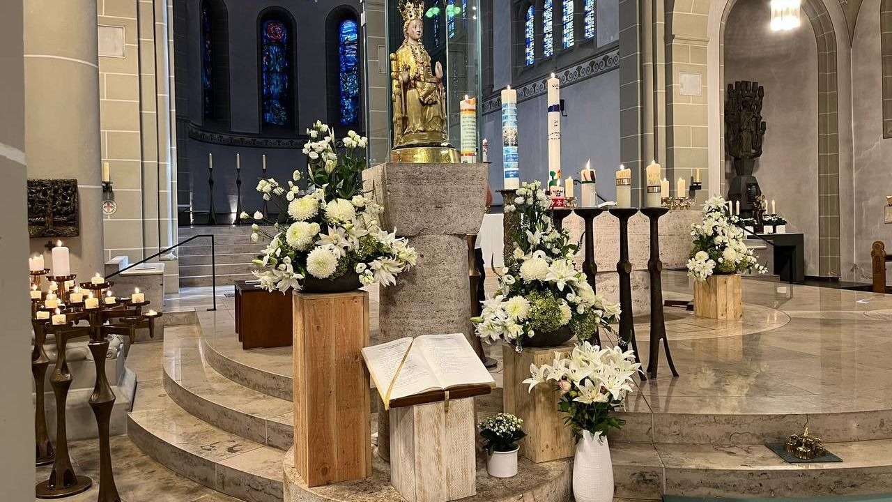 Altarraum einer Kirche mit goldener Marienstatue, weißen Blumen, brennenden Kerzen und einem geöffneten Buch