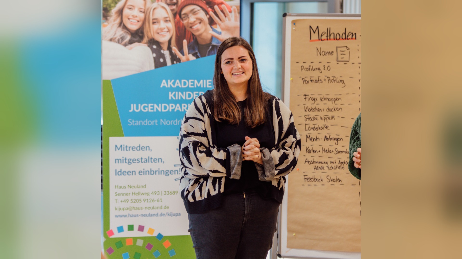 Eine lächelnde junge Frau steht vor einem Banner der Akademie für Kinder- und Jugendparlamente und einem