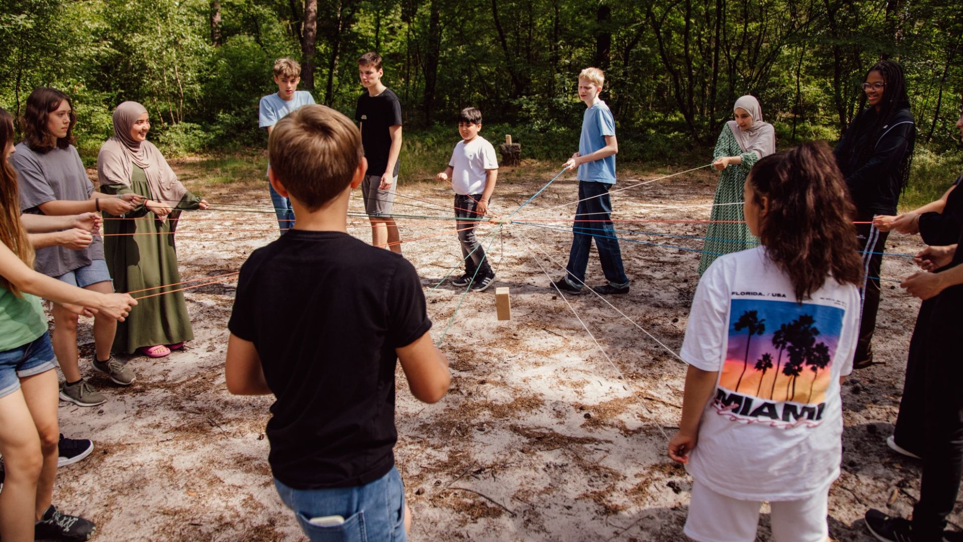 Jugendliche halten im Wald bunte Seile, die zu einem Holzklotz in der Mitte führen, für ein Teamspiel