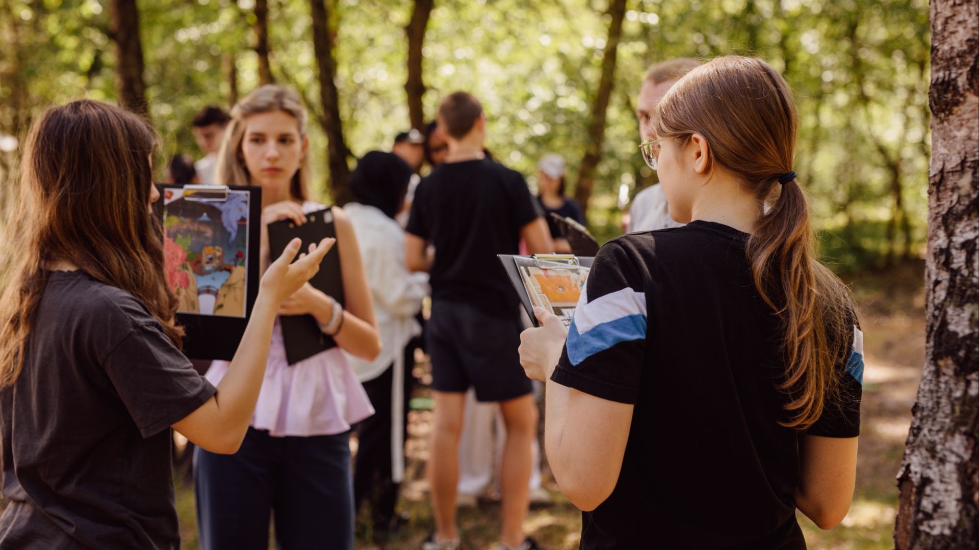 Zwei Jugendliche halten Klemmbretter mit bunten Zeichnungen und sprechen in einer Gruppe im Wald