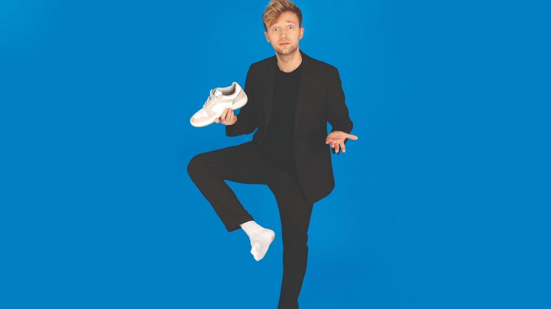 Marc Weide im schwarzen Anzug steht auf einem Bein, hält einen weißen Sneaker und schaut erstaunt vor blauem Hintergrund