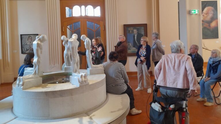 Ältere Besucher betrachten einen großen Marmorbrunnen mit menschlichen Skulpturen in einem Museumssaal mit Gemälden an