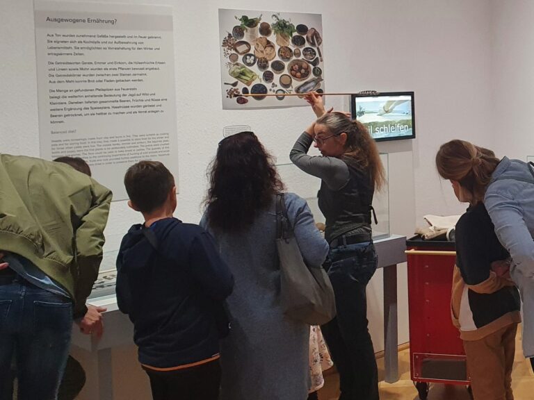 Besucher in einer Ausstellung: Eine Frau erklärt mit einem Stab vor einer Schautafel, andere schauen Displays an