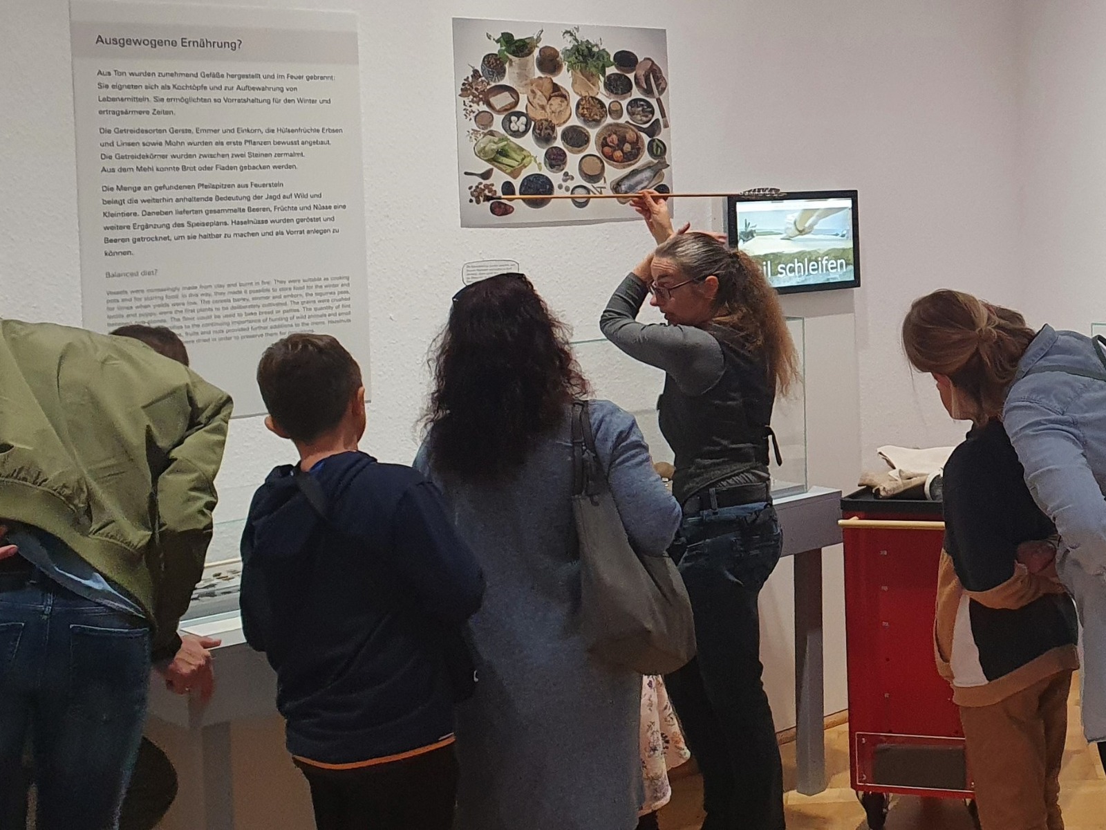 Besucher in einer Ausstellung: Eine Frau erklärt mit einem Stab vor einer Schautafel, andere schauen Displays an