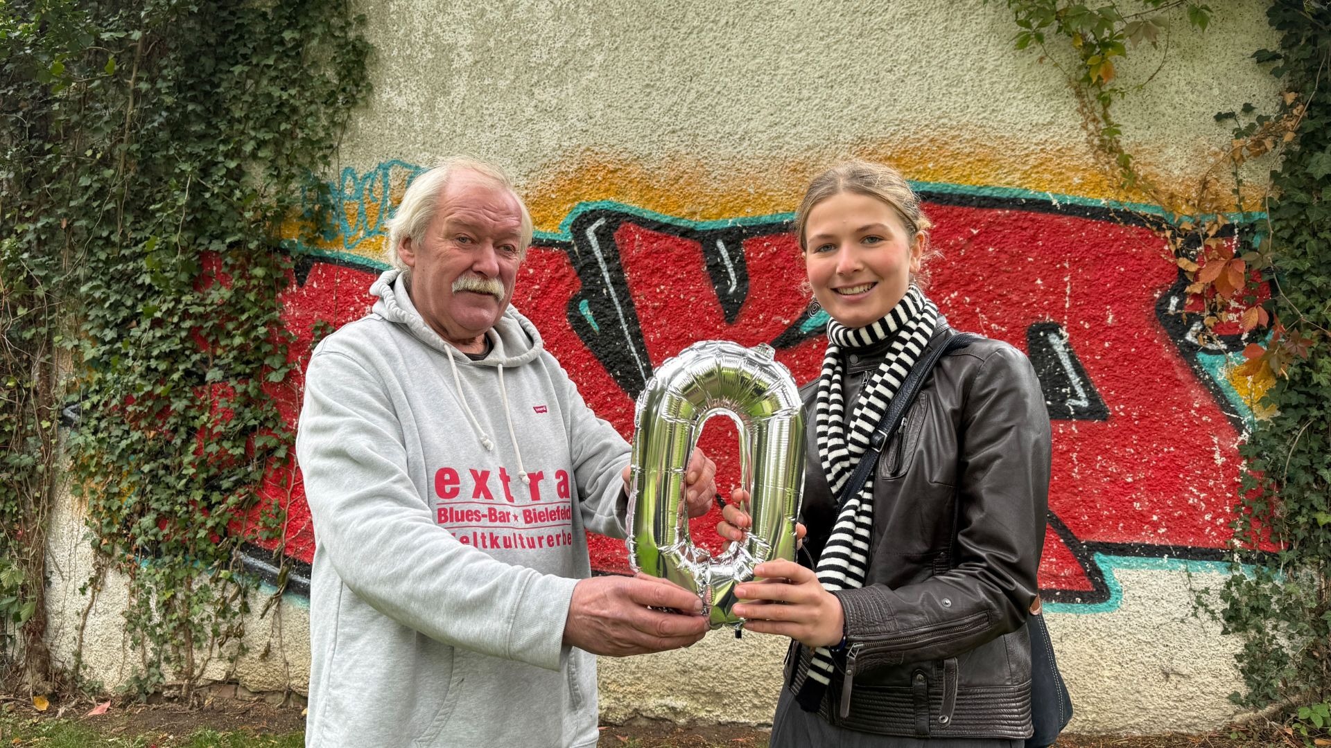 Ein Mann und eine Frau halten gemeinsam einen silbernen „0“-Folienballon vor einer bunten Graffitiwand in Bielefeld