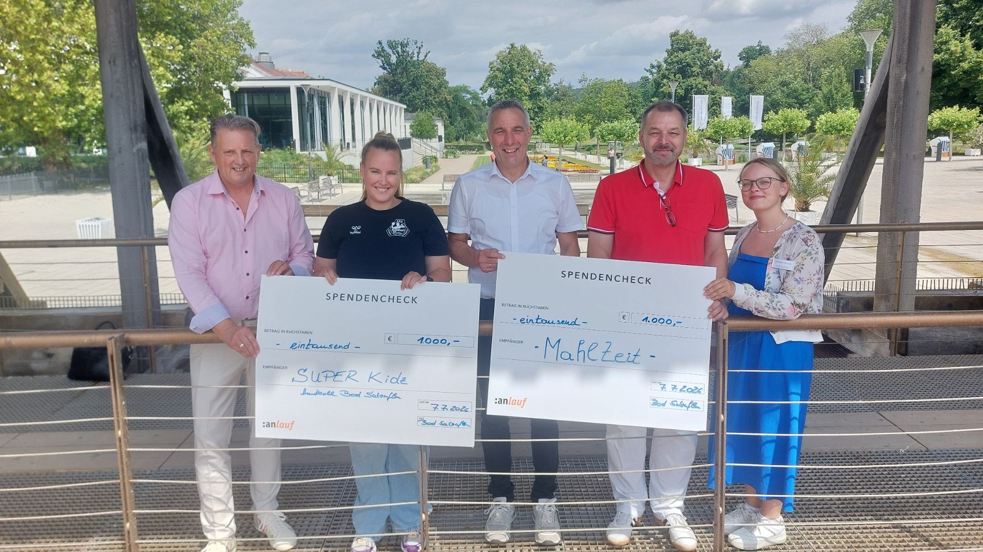 Fünf Personen halten zwei große Spendenschecks über je 1000 Euro für "SUPER Kide" und "MahlZeit" in einer Parkanlage