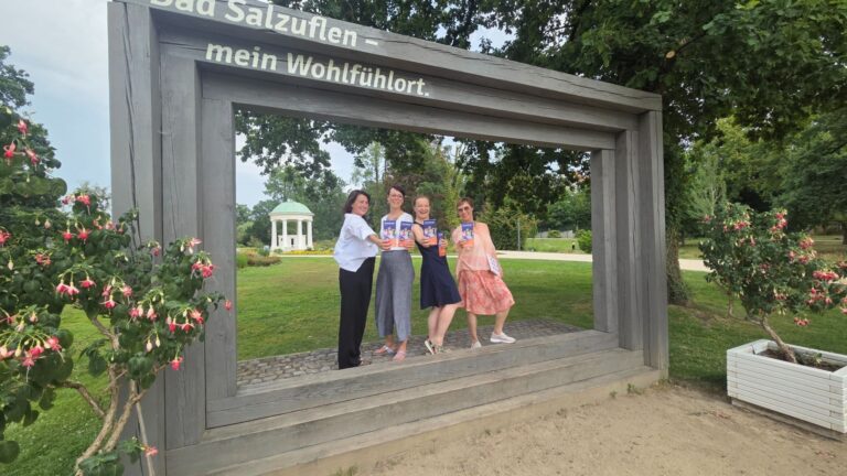 Vier lächelnde Frauen präsentieren Programmhefte in einem großen Holzrahmen mit der Aufschrift Bad Salzuflen in einem