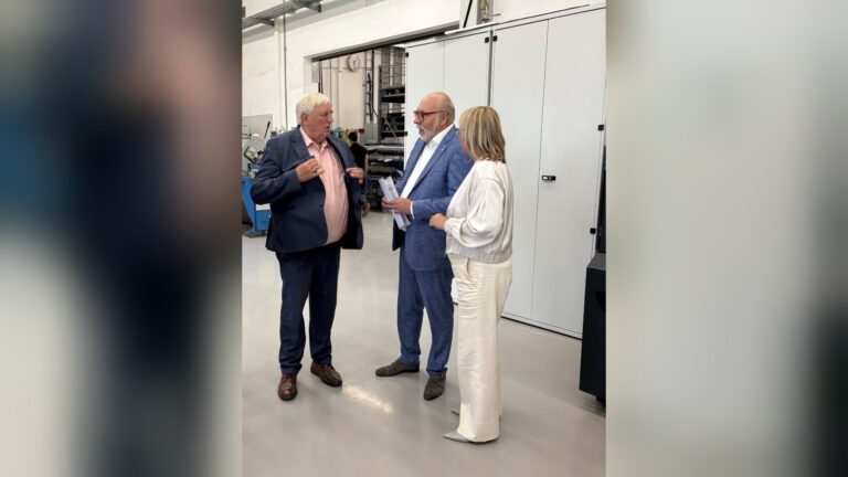 Drei Personen sprechen miteinander in einem Industrieraum vor weißen Schränken, einer hält Dokumente