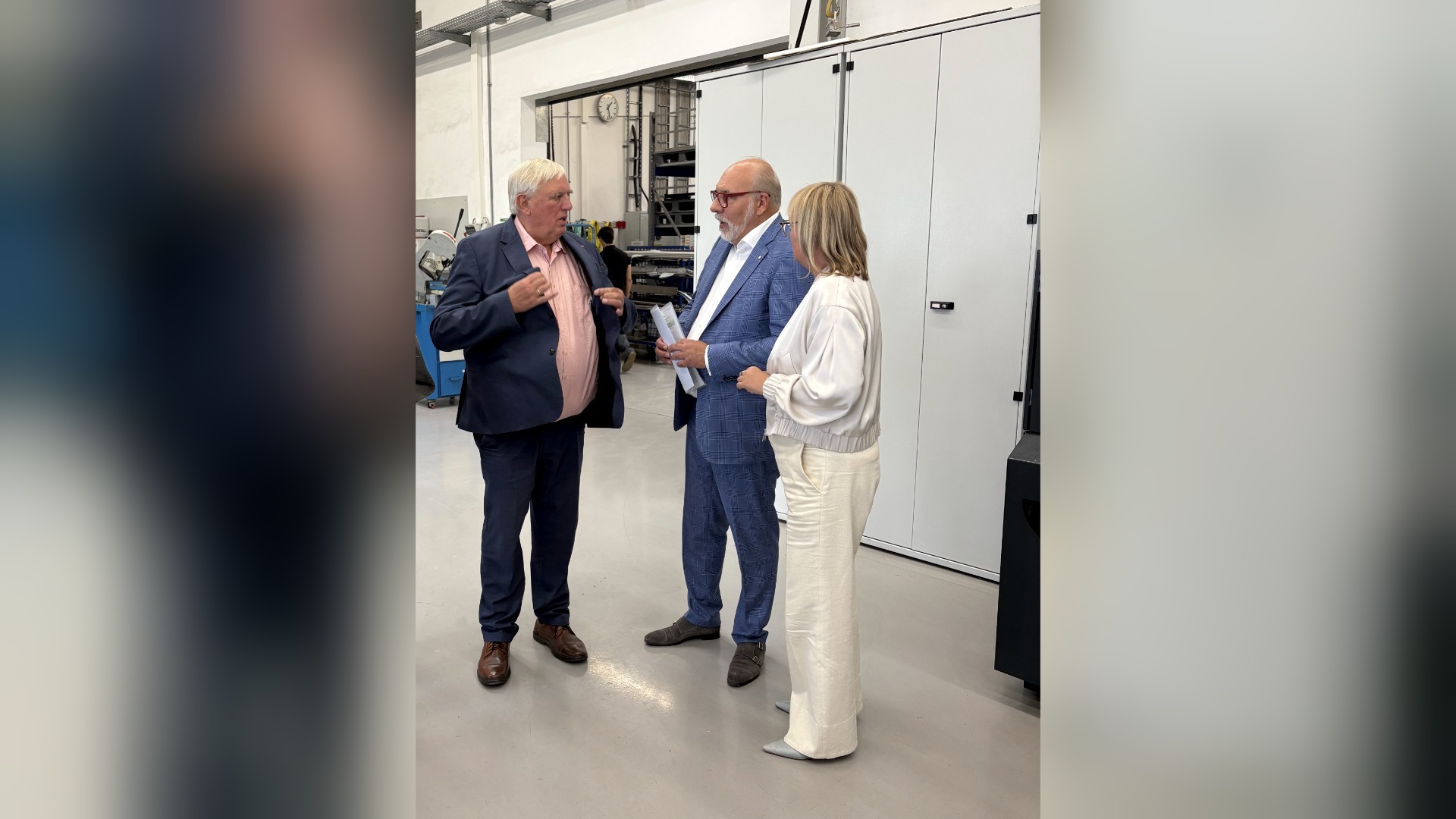 Drei Personen sprechen miteinander in einem Industrieraum vor weißen Schränken, einer hält Dokumente